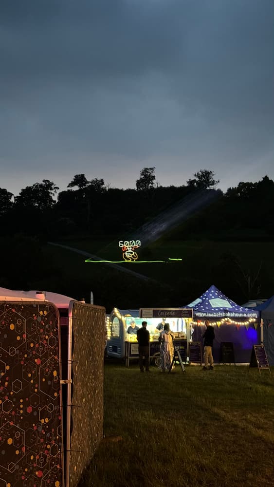 A photo of a hillside at night seen above some food stalls. Projected onto the hillside using lasers is the game "Duck Hunt"